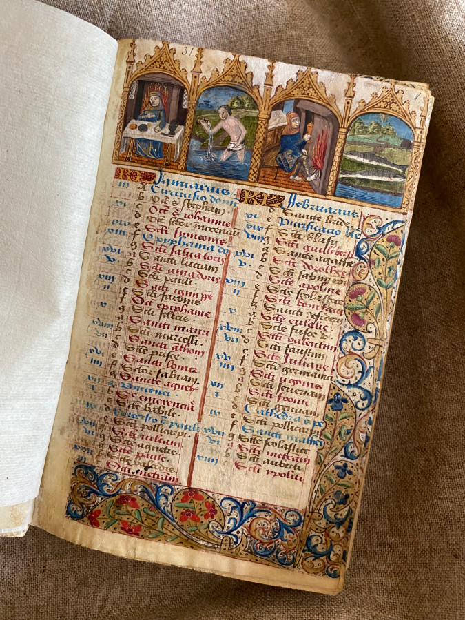 Na grubym płótnie leży otwarta księga Liber precum latinarum et gallicarum cum Calendario. W księdze kolorowe ilustracje, średniowieczny tekst łaciński i ozdobne marginesy. 